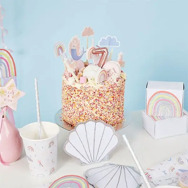 Colorful sprinkle-covered birthday cake topped with pastel rainbow decorations and a number 7 candle, surrounded by matching rainbow-themed party tableware.